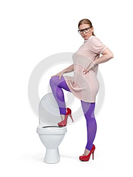 A stylish woman in glasses, a pink dress and red stiletto shoes stands with her foot on the toilet, isolated on a white