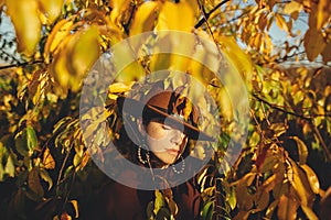 Stylish woman in hat and brown clothes posing among autumn leaves in warm sunny light. Portrait of fashionable young female