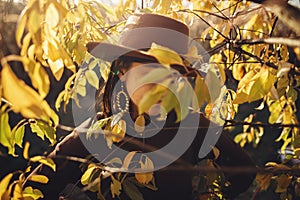 Stylish woman in hat and brown clothes posing among autumn leaves in warm sunny light. Portrait of fashionable young female