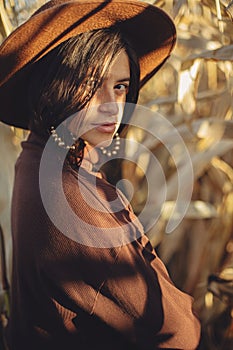 Stylish woman in hat and brown clothes posing in autumn maize field in warm sunny light. Portrait of fashionable attractive young