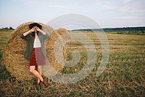 Stylish woman in hat standing at hay bale in summer evening in field. Tranquility. Rural slow life