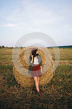 Stylish woman in hat standing at hay bale in summer evening in field. Tranquility. Rural slow life