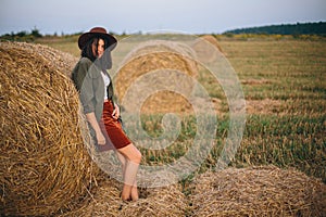 Stylish woman in hat standing at hay bale in summer evening in field. Tranquility. Rural slow life