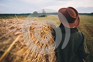 Stylish woman in hat with straw standing at haystack in summer evening countryside field.Tranquility