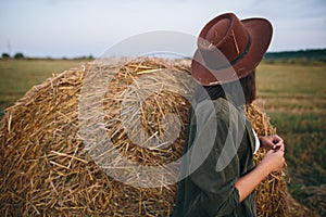 Stylish woman in hat with straw standing at haystack in summer evening countryside field.Tranquility