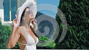 Stylish woman in hat and sunglasses enjoying sunbathing and smiling at summer hotel park