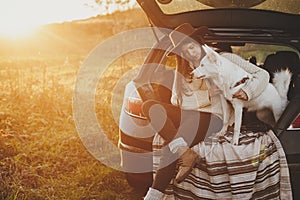 Stylish woman in hat and sweater sitting with cute dog in car trunk in warm sunset light in autumn field. Travel and road trip