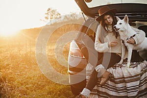 Stylish woman in hat and sweater sitting with cute dog in car trunk in warm sunset light in autumn field. Travel and road trip