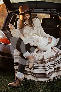 Stylish woman in hat and sweater sitting and hugging cute dog in car trunk in evening autumn field. Road trip with pet.Young