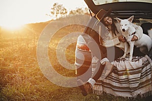 Stylish woman in hat and sweater sitting and hugging cute dog in car trunk in warm sunset light in autumn field. Road trip with