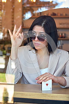 Stylish woman with a long manicure, fashionable sunglasses and a colored jacket through the glass of a cafe.