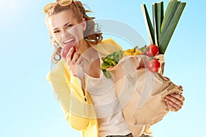 Stylish woman with paper bag with groceries biting apple a