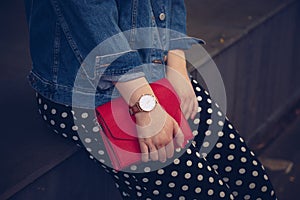 Stylish woman in polka dot culottes and denim jacket holding a red purse and wearing a rose gold wrist watch.