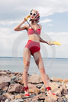 stylish woman in red bikini and silk scarf talking on yellow rotary phone and standing