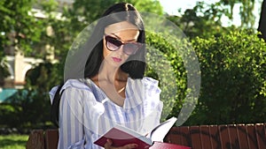 Stylish woman in sunglasses and stripped blouse read book in summer park