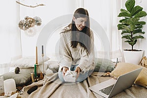 Stylish woman in sweater holding furoshiki gift in festive decorated room and looking at laptop