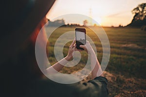 Stylish woman taking photo of sunset on phone in summer field. Atmospheric beautiful moment