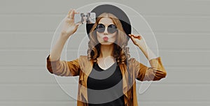 Stylish woman taking selfie picture by smartphone wearing black hat, sunglasses, brown jacket with curly hair over gray