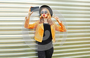 Stylish woman taking selfie picture by smartphone wearing yellow jacket, black round hat on metal wall