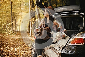 Stylish woman traveller with backpack sitting with cute dog in car trunk in sunny autumn woods. Young hipster female traveling