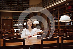 Stylish woman in a white blouse sitting at a long table in an old public library and reading a book with a serious face