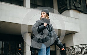 Stylish woman in a winter jacket and hat walks down the street with a shopper bag in his hands on the background of the building