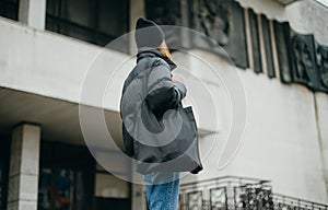 Stylish woman in a winter jacket and hat walks down the street with a shopper bag in his hands on the background of the building