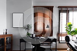 Stylish working corner decorate with black wood empty frame and artificial plant in glass vase and gold mirror stationary in