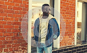 Stylish young african man model wearing denim jacket posing on city street over brick wall background