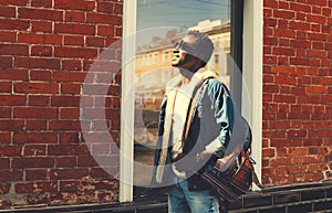 Stylish young african man wearing jeans jacket and backpack looking up at sunlight while walking on city street over brick wall