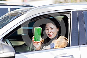 Stylish young asian woman sitting in car and showing her mobile phone with chromakey green screen. Automobile