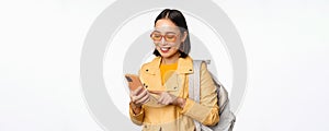 Stylish young asian woman tourist, traveller with backpack and smartphone smiling at camera, posing against white