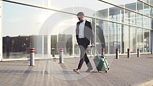 Stylish young bearded man in sunglasses exiting the airport terminal with luggage, answers the phone. Business style