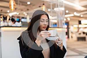 Stylish young beautiful brunette woman with long hair in black fashionable coat with cup of coffee is resting in cafe in mall.