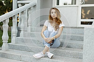 Stylish young beautiful woman in trendy summer T-shirt in fashionable jeans in trendy sneakers resting sitting on the steps