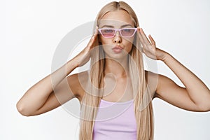 Stylish young blond woman wearing trendy sunglasses, pucker lips and looking sassy at camera, standing over white