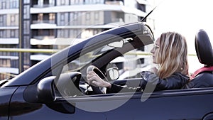 Stylish, young blonde woman driving her beatiful black sportish convertible car. Side, slow motion footage. Lean a hand