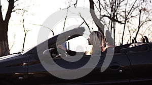 Stylish, young blonde woman driving her beatiful black sportish convertible car. Side, slow motion footage. Silhouettes