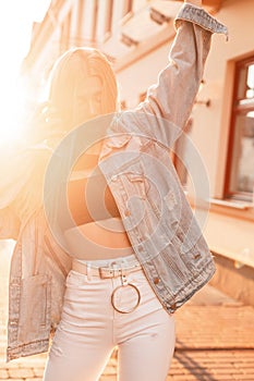 Stylish young blonde woman in fashionable denim clothes posing on the street against the backdrop of bright sunlight at sunset