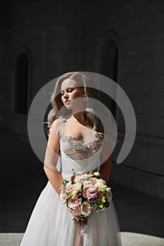 Stylish young bride in classical wedding dress posing with a bridal bouquet outdoors. Female model in luxurious wedding