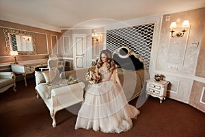 A stylish young bride wearing luxurious wedding dress sits on the bed in a vintage interior. Beautiful model girl with