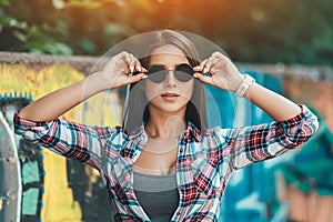 Stylish young brunette woman on graffiti background. Portrait of a fashionable female in sunglasses. Sunlight background