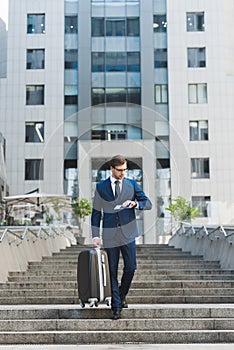 stylish young businessman with luggage and flight tickets walking by business district and looking