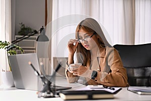 Stylish young businesswoman wearing glasses drinking coffee and reading email on her laptop computer