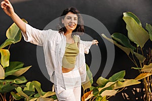 Stylish young caucasian girl enjoys free time standing near black wall with plants.