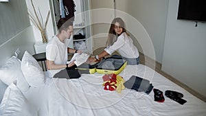 Stylish young couple sitting on bed with opened suitcase and packing up things for travel. Man and woman thinking about