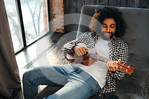 Stylish young egyptian man lies on the sofa and plays the guitar against the background of a large window