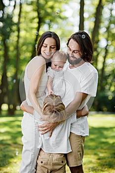 Stylish young family is having rest in the park. Dad and mom are holding daughter in the arms and hugging son.