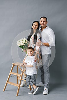 Stylish young family: mom, dad and four-year-old son stand near a wooden stepladder and smile, isolated on a white background.