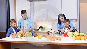 Stylish young family with two sons preparing healthy vegetarian breakfast with fresh vegetables on cozy home kitchen.